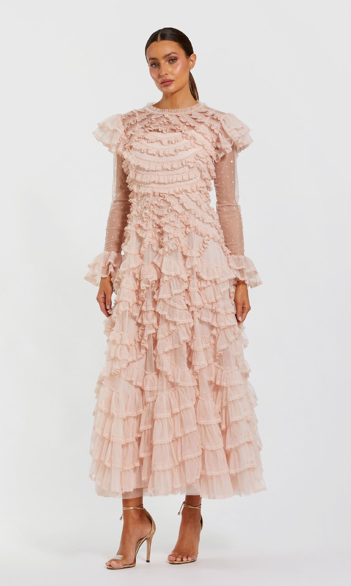 A woman models the Mac Duggal 8274 Long Sleeve Ruffle Formal Dress in light pink with sheer sleeves and nude high-heeled sandals, posing against a plain white background.
